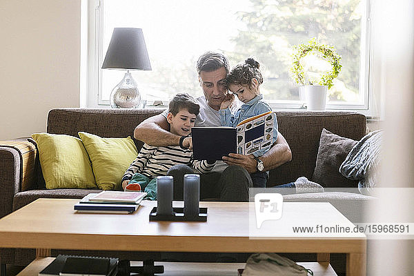 Vater liest Sohn und Tochter ein Buch vor  während sie zu Hause auf dem Sofa sitzen