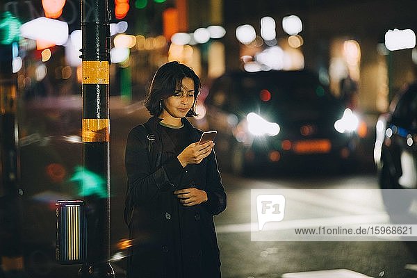 Woman text messaging through smart phone while standing in city at night