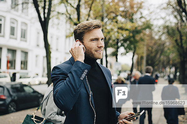 Geschäftsmann schaut weg  während er in der Stadt In-Ear-Kopfhörer hält