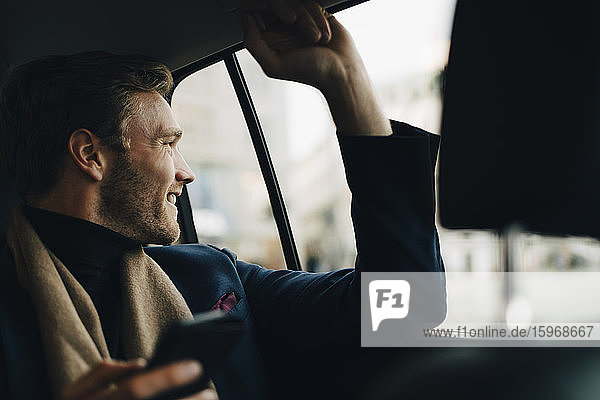 Lächelnder Geschäftsmann mit Telefon  der durchs Fenster schaut  während er im Taxi sitzt