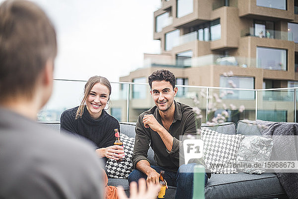 Smiling man and woman looking at female friend while sitting on sofa during party at building terrace