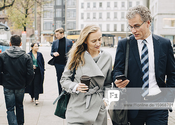 Businessman showing smart phone to female coworker while walking in city