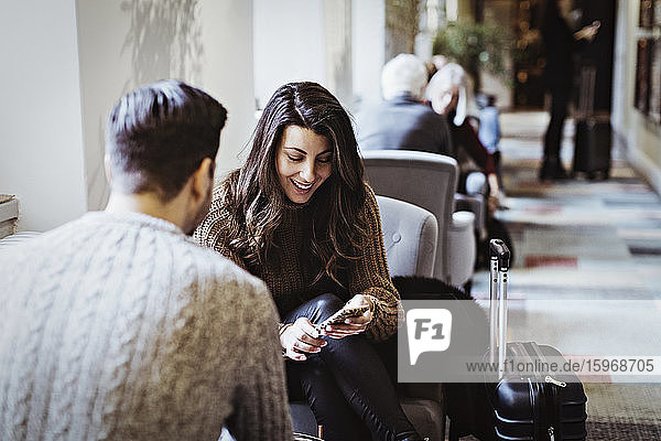 Lächelnde Frau benutzt Smartphone  während sie mit ihrem männlichen Partner auf einem Stuhl im Hotel sitzt