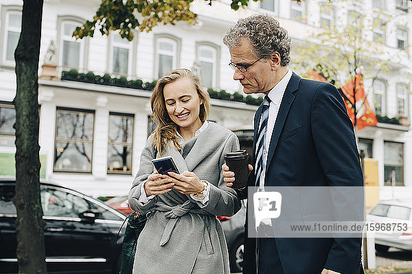 Lächelnde Geschäftsfrau zeigt einem männlichen Kollegen in der Stadt ein Smartphone