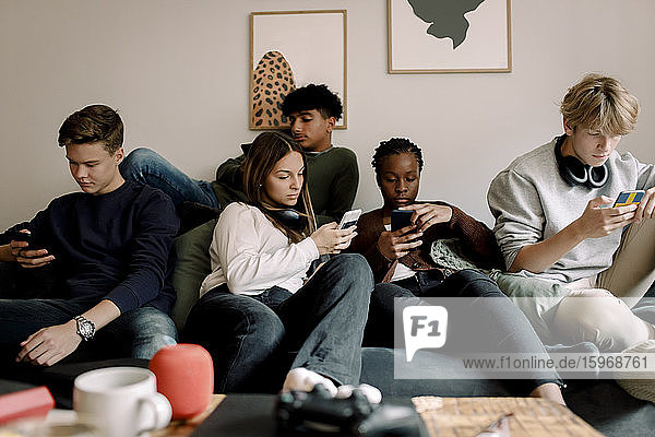 Multi-ethnische Freunde nutzen soziale Medien auf Mobiltelefonen  während sie zu Hause auf dem Sofa sitzen