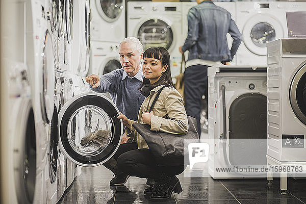 Reifere Besitzerin zeigt auf Waschmaschine  während weibliche Kundin im Elektronikgeschäft kauert