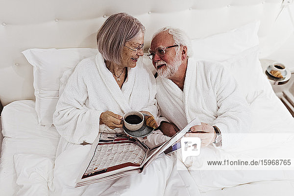 Ältere Frau sieht Mann mit Zeitung in der Hand an  während sie im Hotel Kaffee auf dem Bett trinkt