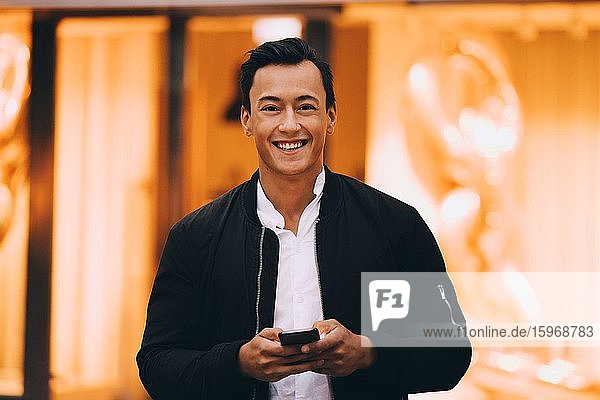 Porträt eines lächelnden Mannes  der ein Mobiltelefon benutzt  während er in einer beleuchteten Stadt steht