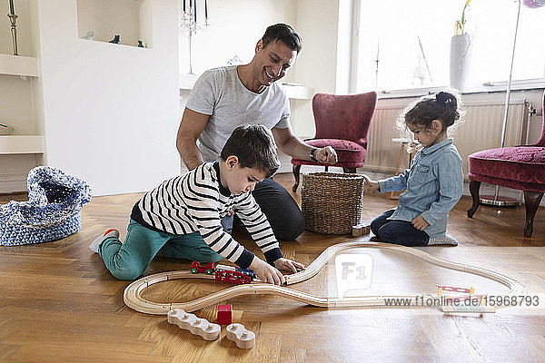Sohn spielt mit Miniatur-Eisenbahn  während Vater und Schwester zu Hause lächelnd auf dem Boden knien