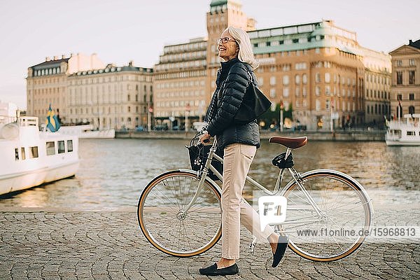 Side view of smiling senior woman with bicycle looking up while crossing river in city