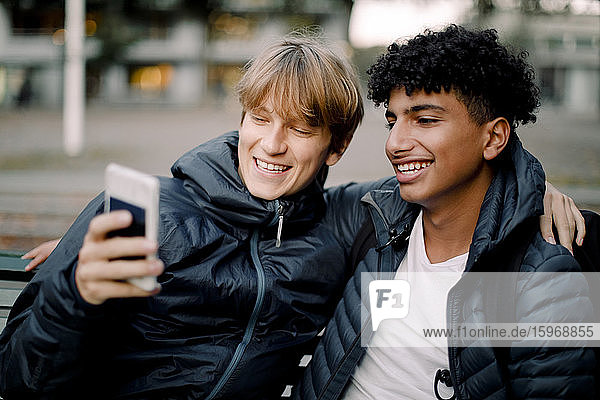 Smiling teenage boy taking selfie through smart phone while sitting with friend in city