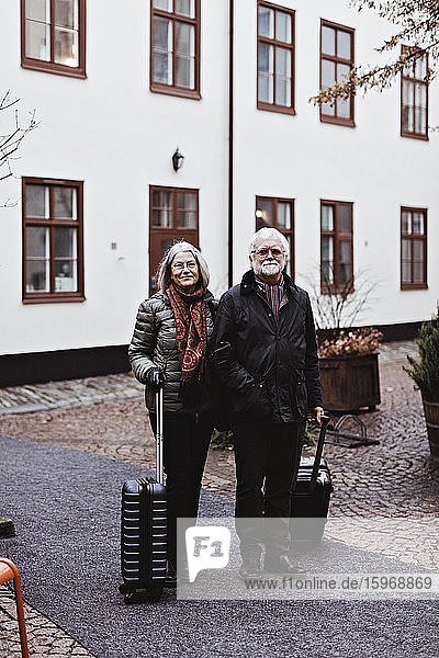 Portrait of senior couple standing on footpath with luggage against hotel