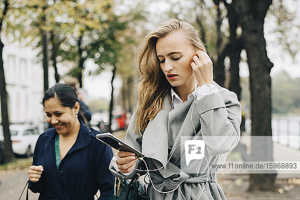 Businesswoman wearing in-ear headphones while looking at smart phone in city