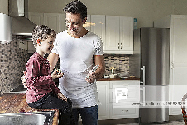 Smiling father showing smart phone to son with food while standing by kitchen counter