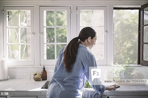 Rückansicht einer Geschäftsfrau  die ein Smartphone auf der Küchentheke im Heimbüro benutzt
