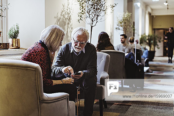 Älterer Mann zeigt einer Frau ein Smartphone  während er im Hotel auf einem Stuhl sitzt