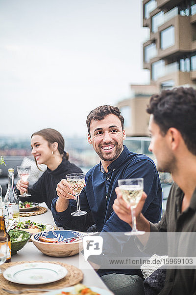 Lächelnder Mann unterhält sich mit einem Freund  während er während einer Party auf dem Dach neben einer Frau sitzt