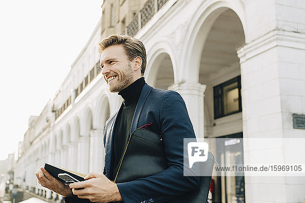 Lächelnder Geschäftsmann mit Smartphone schaut weg  während er gegen ein Gebäude in der Stadt steht