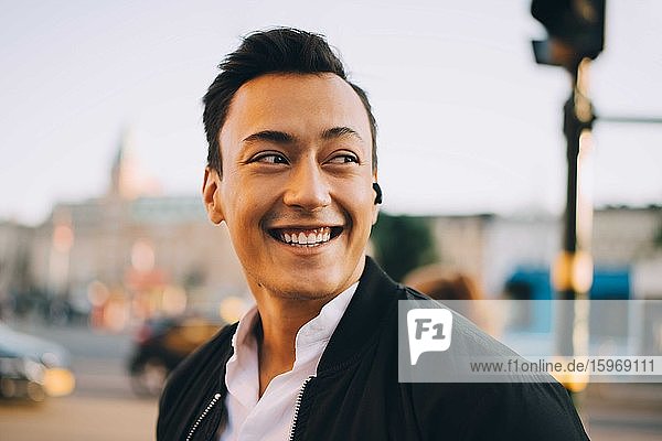 Lächelnder Mann schaut weg  während er in der Stadt In-Ohr-Kopfhörer trägt