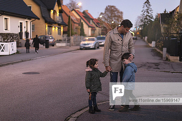 Vater tröstet traurigen Sohn  während er mit der Tochter bei Sonnenuntergang auf dem Fußweg Händchen hält