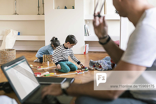 Vater mit Laptop schaut Kindern beim Spielen zu Hause zu