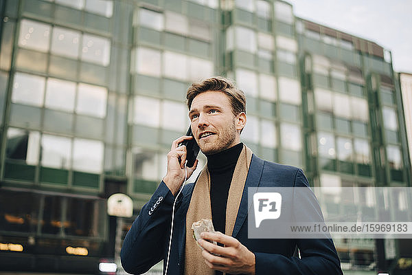 Selbstbewusster Geschäftsmann mit Wrap-Sandwich  der in der Stadt stehend per Smartphone spricht