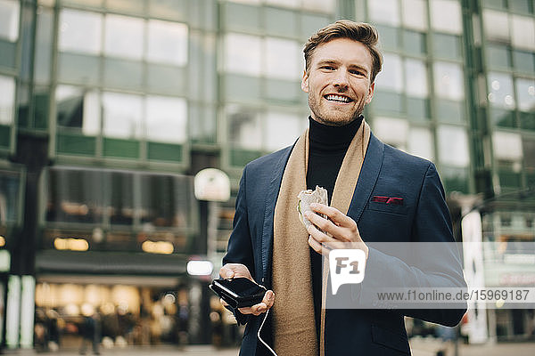 Portrait of smiling businessman with wrap sandwich standing in city
