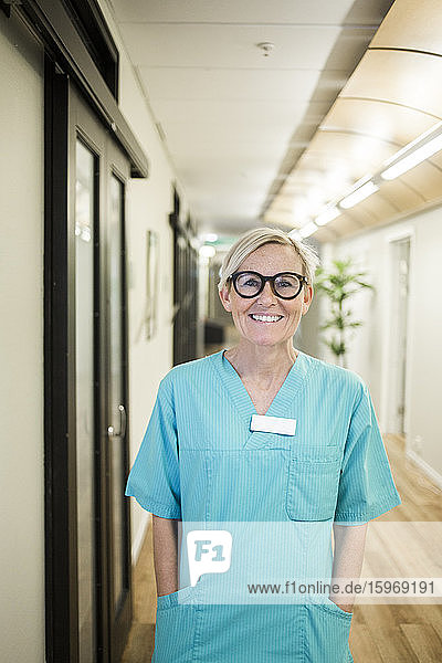 Porträt einer reifen Mitarbeiterin im Gesundheitswesen mit Händen in den Taschen im Krankenhauskorridor stehend