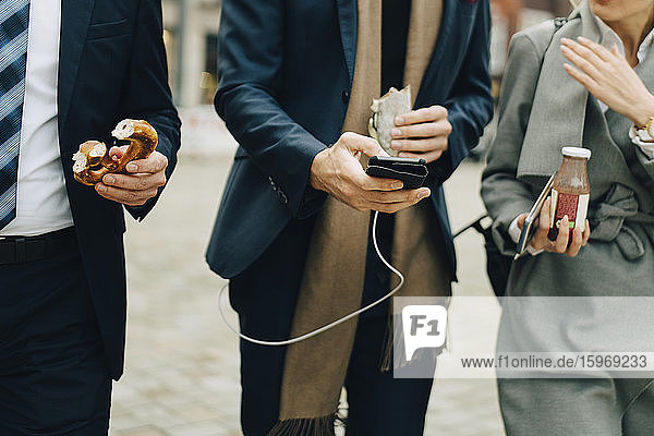 Ein Teil der Geschäftsleute hält Brezeln und Getränke in der Hand  während sie ein Smartphone in der Stadt benutzen