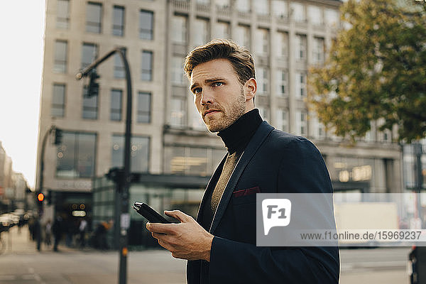 Contemplating businessman with smart phone looking away while standing against building in city