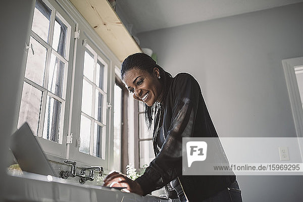 Lächelnde weibliche Fachkraft schaut auf den Laptop  während sie im Heimbüro steht