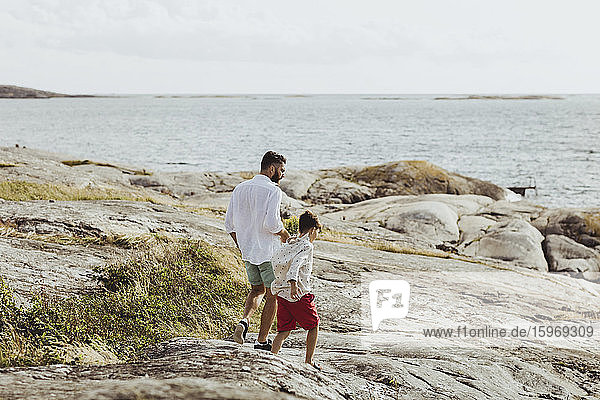 Rear view of father and son walking towards sea on rocky land
