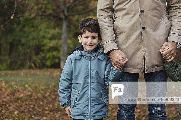Porträt eines lächelnden Sohnes  der im Herbst mit dem Vater Händchen hält  während er im Park steht