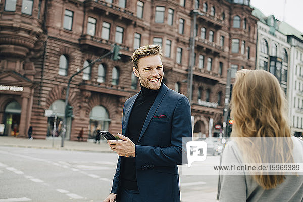Lächelnder Geschäftsmann mit Telefon  der eine in der Stadt stehende Mitarbeiterin ansieht