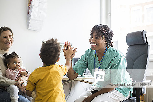 Lächelnder Kinderarzt gibt dem Jungen High-Five  während die Mutter in der Klinik liebevoll hinsieht