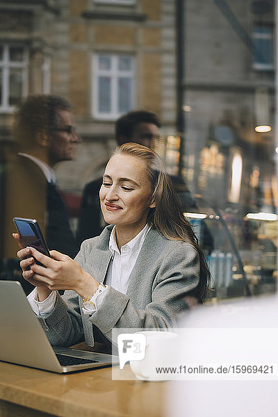 Lächelnde Geschäftsfrau mit Smartphone im Café durch Glasfenster gesehen