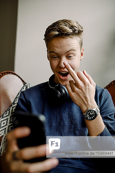 Teenage boy showing middle finger while using smart phone at home