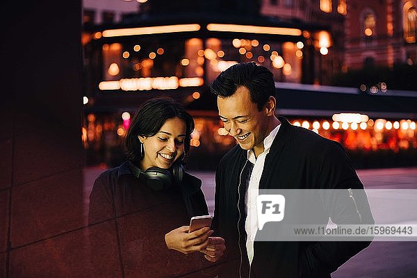Lächelnder Mann und lächelnde Frau  die ein Smartphone benutzen  während sie nachts in der Stadt stehen