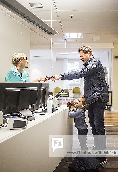 Side view of mature man giving id card to doctor while son standing by reception in hospital