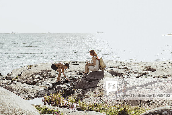 Seitenansicht einer Frau  die sitzt  während ihr Sohn sich bei Sonnenschein gegen das Meer beugt
