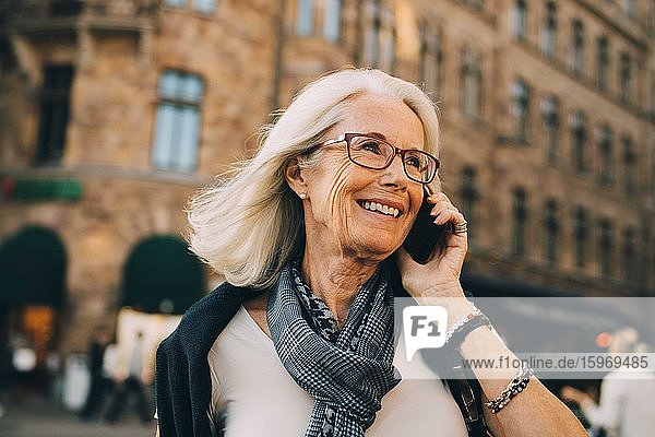 Smiling wrinkled woman talking through phone while looking away in city