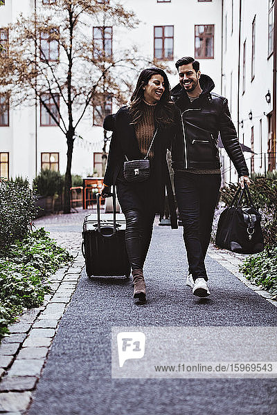 Full length of happy couple walking on footpath with luggage outside hotel