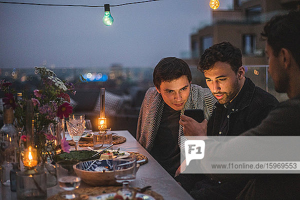 Mann zeigt männlichen Freunden ein Smartphone  während er bei geselligem Beisammensein auf der Terrasse sitzt