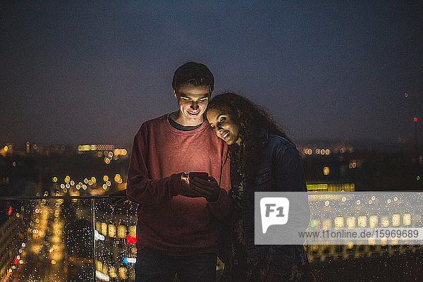Junger Mann zeigt einer Freundin ein Smartphone während eines geselligen Beisammenseins auf einer Terrasse in der Nacht