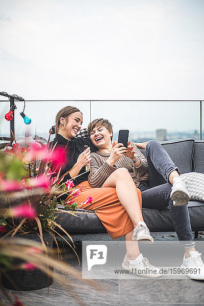 Fröhliche Frau benutzt Smartphone  während sie beim geselligen Beisammensein mit einem fröhlichen Freund auf dem Sofa sitzt