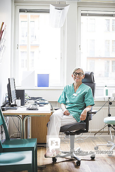 Porträt einer lächelnden  reifen Ärztin in voller Länge  die in der Klinik sitzt