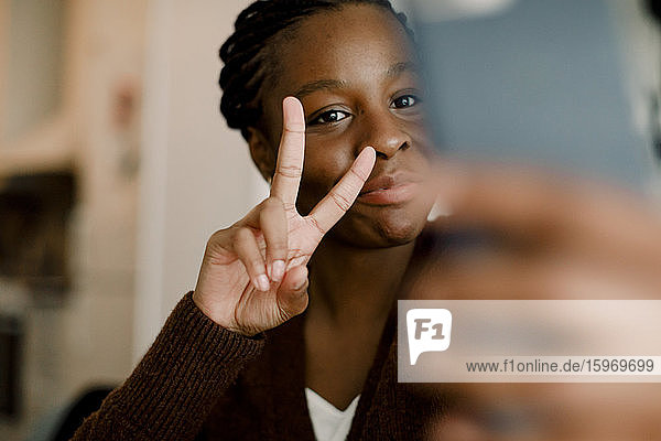 Teenager-Mädchen zeigt Friedenszeichen  während sie sich zu Hause per Smartphone selbstständig macht