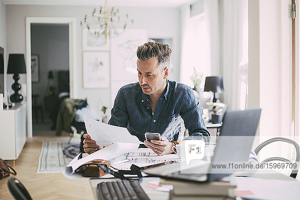 Male architect sitting at workplace reading blueprint at home
