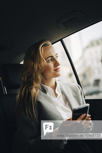 Selbstbewusste Unternehmerin mit Smartphone  die im Taxi sitzend durchs Fenster schaut