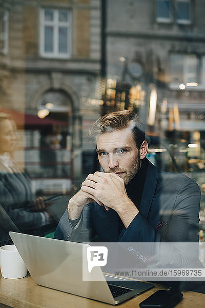 Portrait of businessman with laptop in cafe seen through glass window